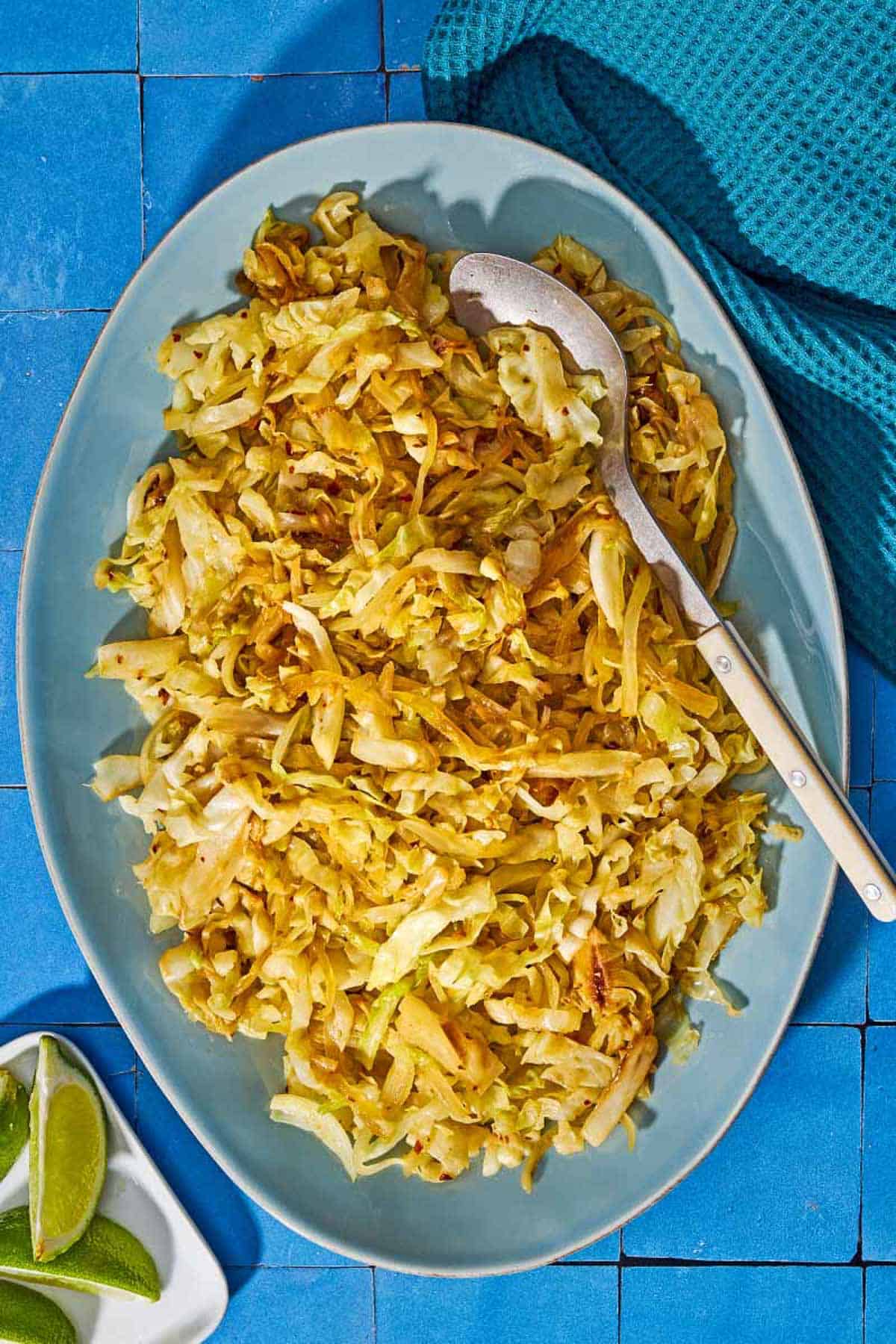 Sauteed cabbage on a platter with a spoon with a plate of lime wedges nearby.