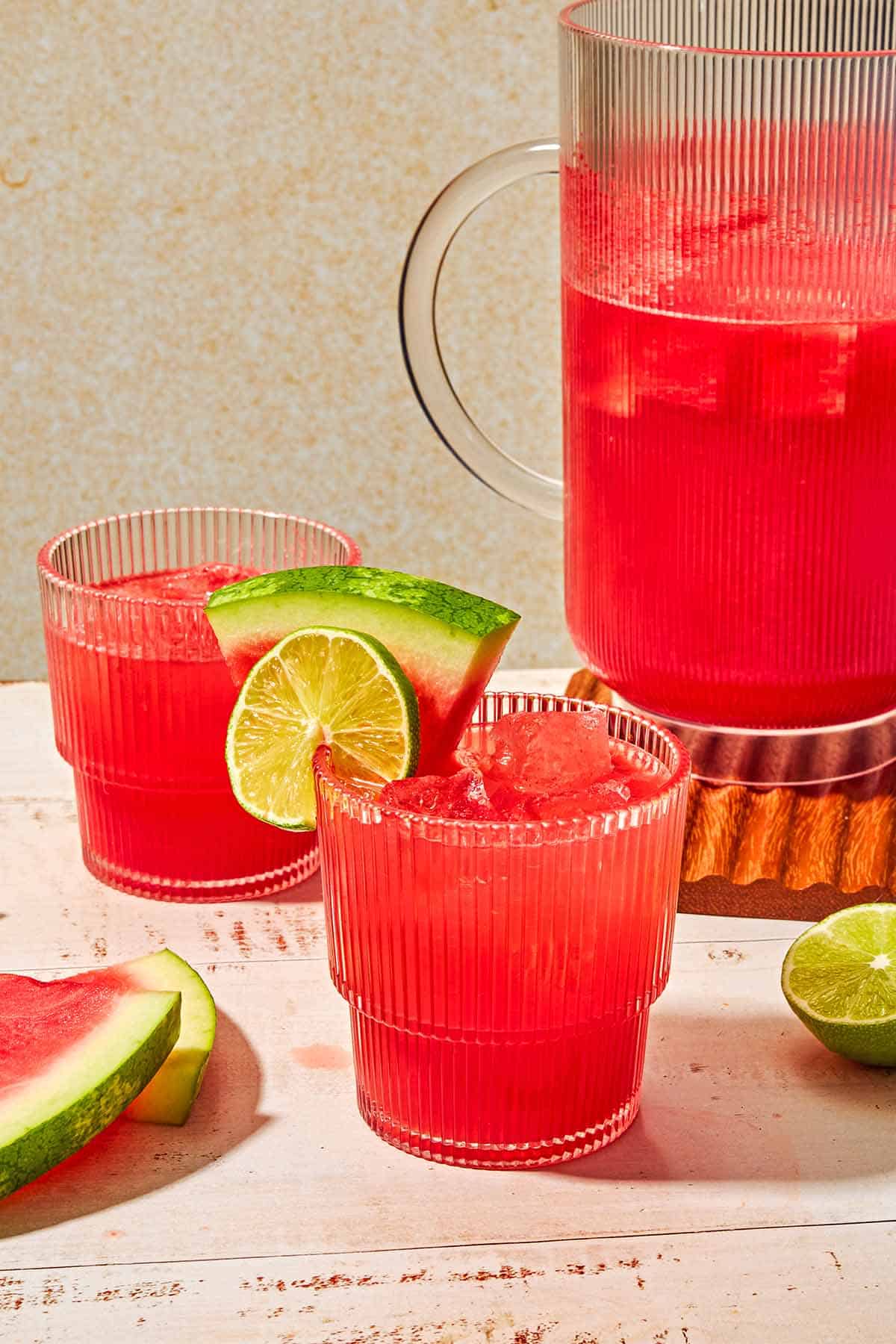 A pitcher of watermelon sangria next to a lime half, 2 watermelon slices, 2 glasses of the sangria, with one garnished with a watermelon slices and a lime wheel.