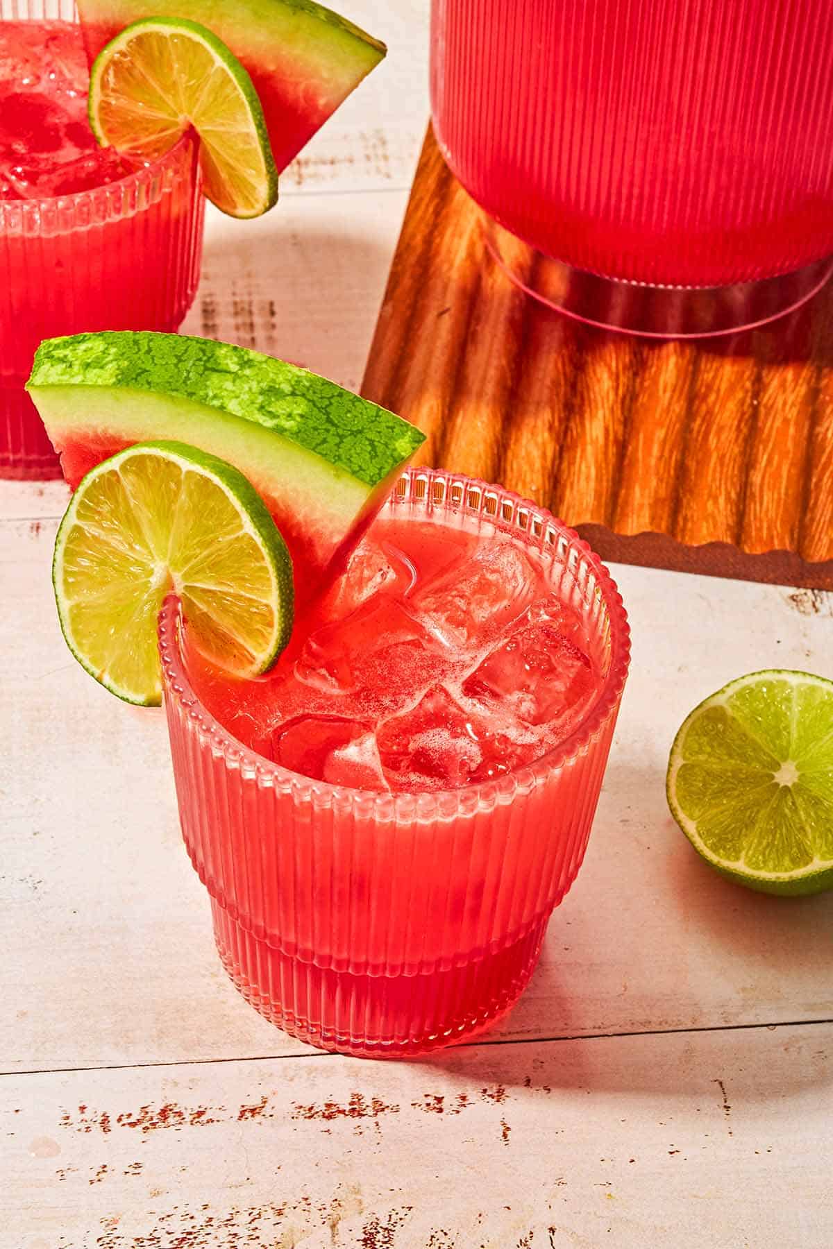 A glass of watermelon sangria garnished with a lime wheel and a watermelon slice next to another glass of the sangria, a pitcher of the sangria, and a lime half.