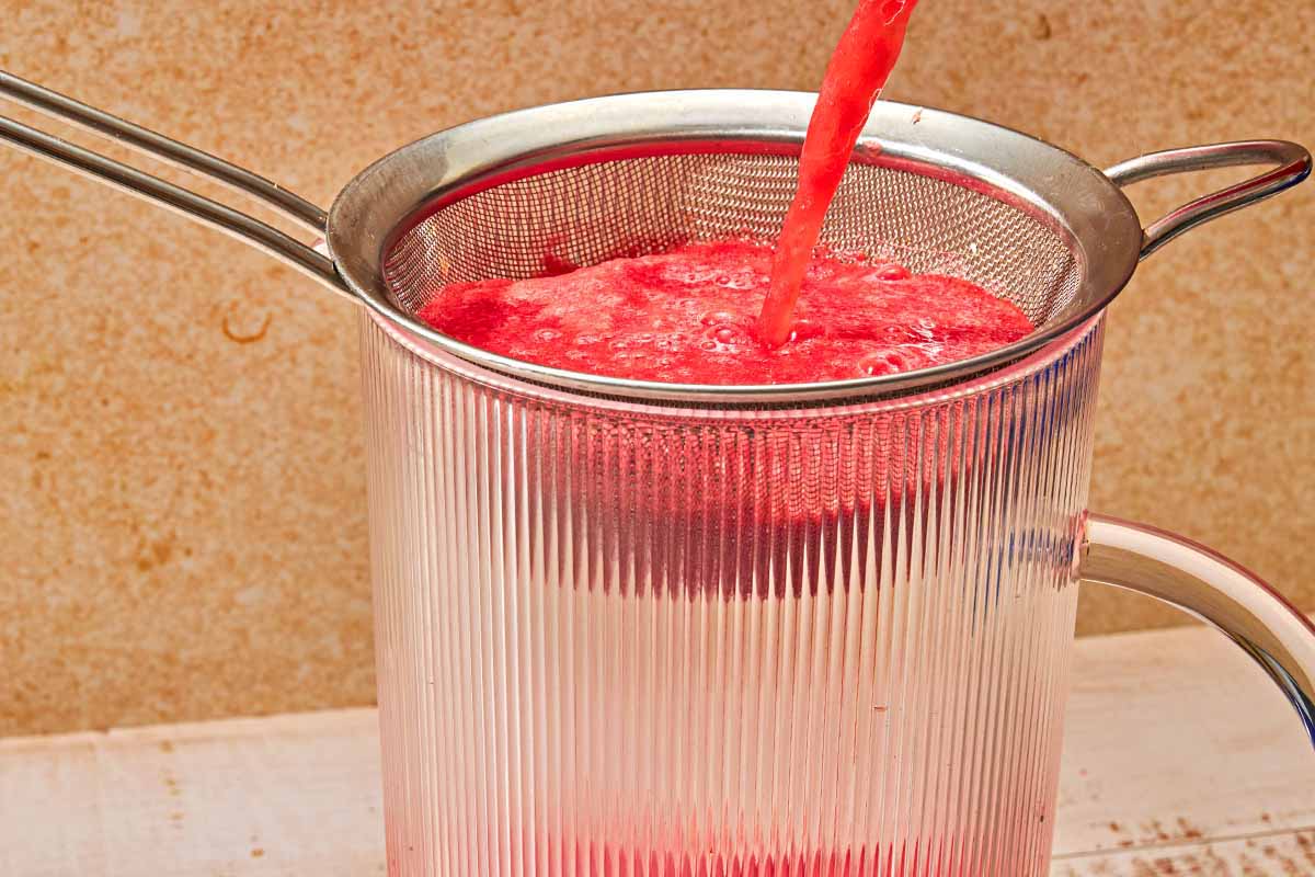 The watermelon sangria being poured through a mesh strainer into a glass pitcher.
