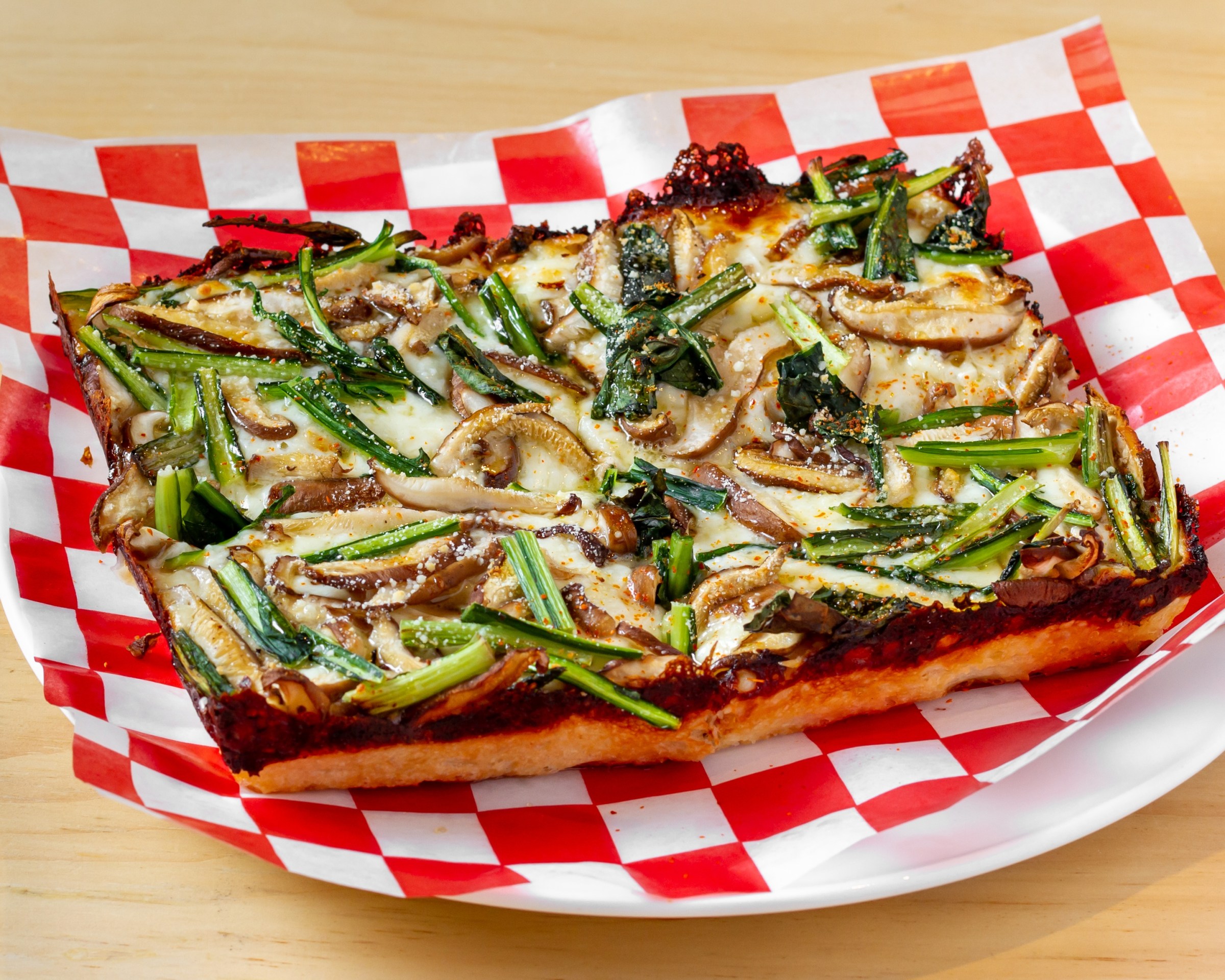 a rectangle of thick detroit-style pizza sits on a plate on top of red-and-white checkered wax paper. the pizza is topped with mushrooms and crispy browned cheese can be seen around the edges of the pizza.