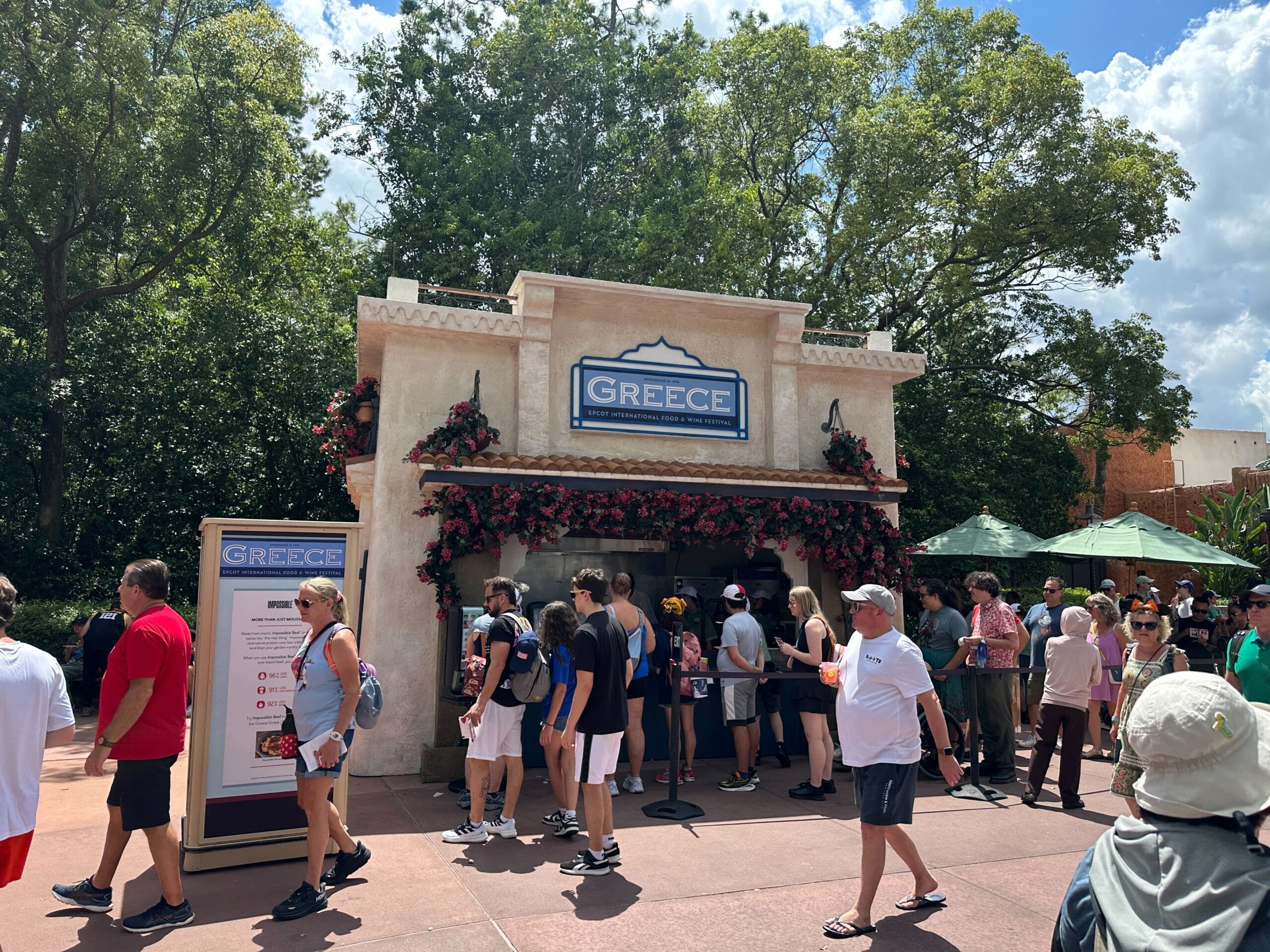 People line up at an outdoor Greece Food & Wine booth surrounded by trees on a sunny day.