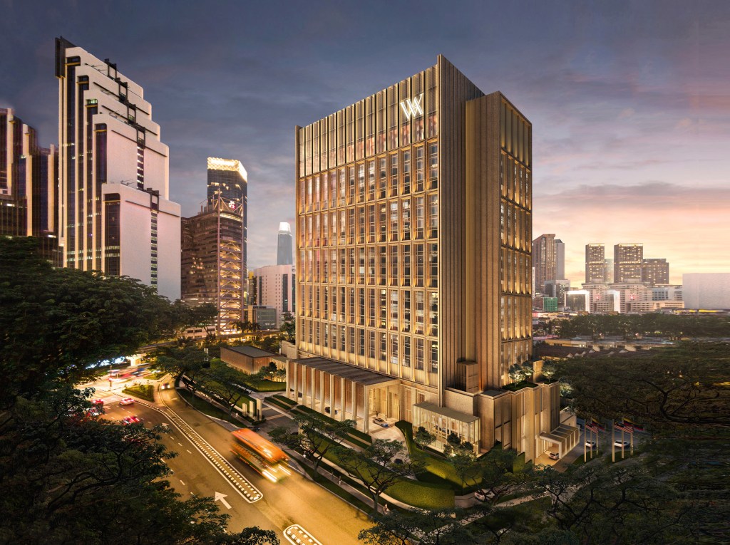Modern office tower with golden lighting at dusk, featuring illuminated “W” logo, city skyline, trees, and traffic on a curved road below.
