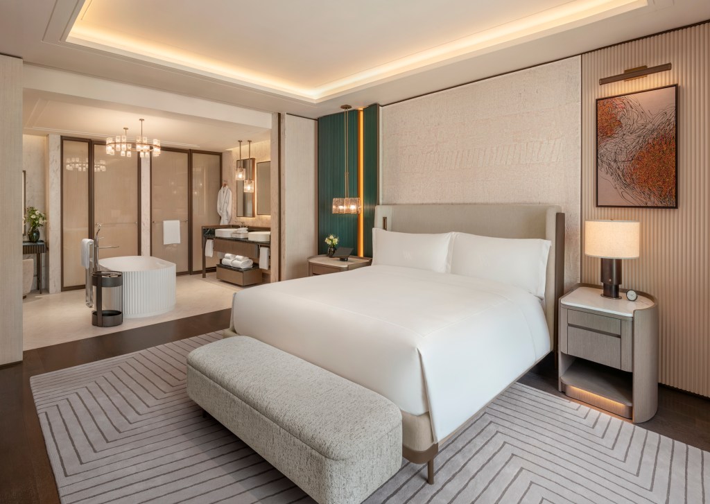 Modern hotel bedroom with white-linen bed, bench, nightstands, geometric rug, abstract art, and open bathroom with freestanding tub and double sinks.