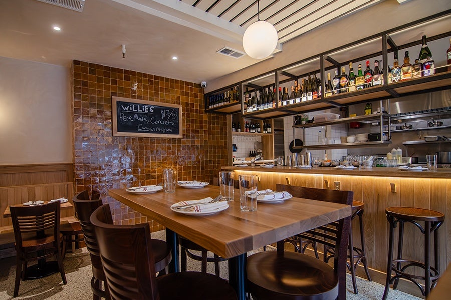 Wooden dining table set with plates, napkins, and glasses in a cozy restaurant interior. The background features a tiled wall with a chalkboard menu, hanging shelves stocked with bottles, and a bar counter with stools. Warm lighting enhances the inviting atmosphere.