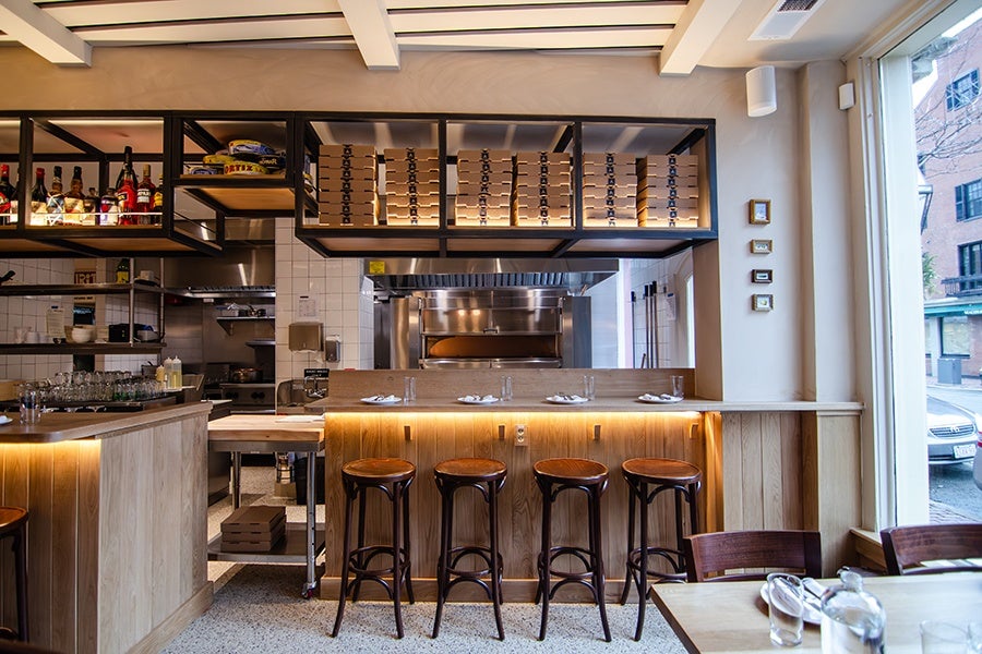Modern restaurant interior featuring a wooden bar counter with four high stools, set with plates and glasses. Above the counter, shelves hold stacked pizza boxes and bottles of liquor. The background includes a pizza oven and kitchen area with stainless steel appliances. Large window on the right lets in natural light.
