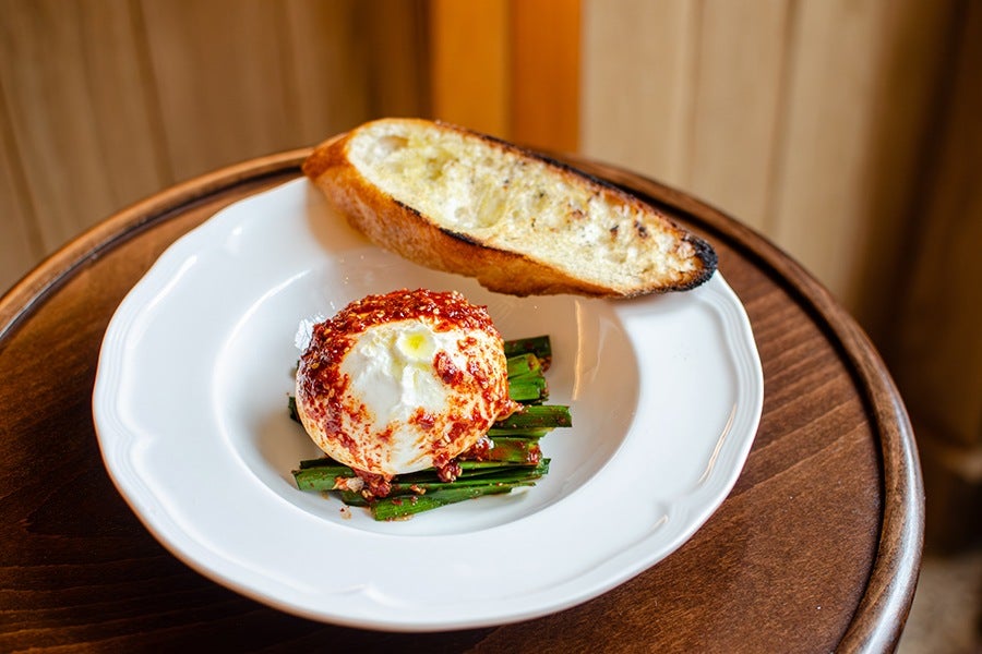 A white plate with a serving of soft cheese topped with red chili flakes, placed on a bed of green vegetables, accompanied by a slice of toasted bread with a golden crust. The plate is set on a round wooden table.