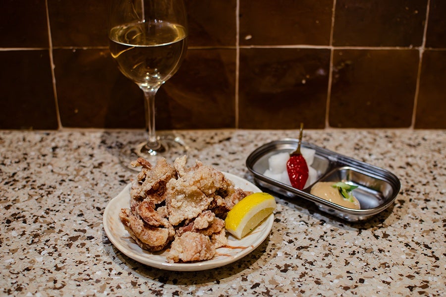 A plate of crispy fried chicken pieces with a lemon wedge, accompanied by a small metal tray containing pickled radish cubes, a red chili pepper, and a dipping sauce garnished with a green herb. A glass of white wine is placed behind the plate on a speckled countertop with brown tiled walls in the background.