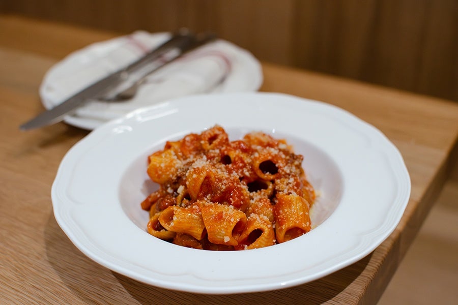 Rigatoni pasta coated in tomato sauce and sprinkled with grated cheese, served in a white bowl on a wooden table. A plate with a knife and fork wrapped in a napkin is in the background.