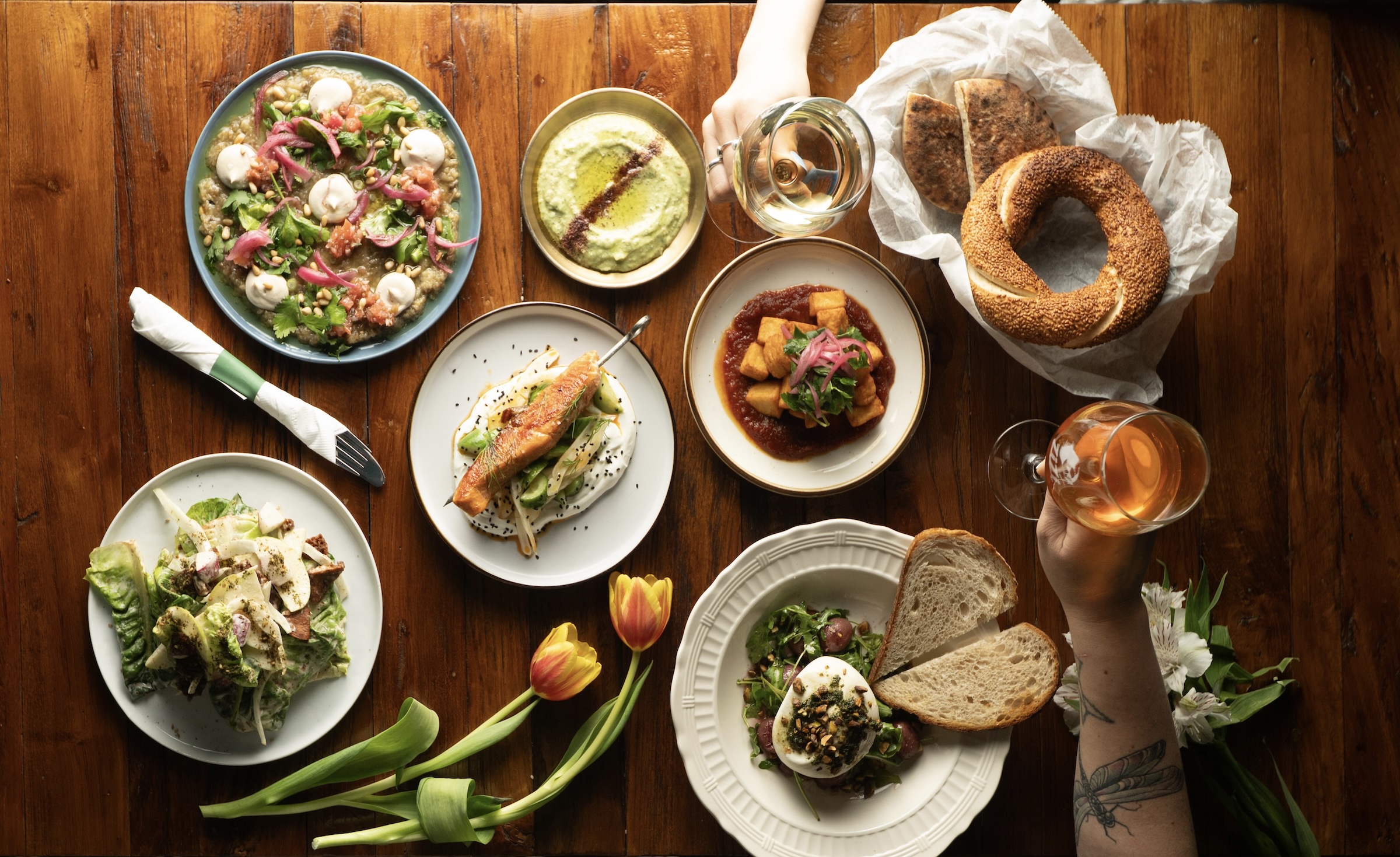 Overhead view of a rustic wooden table filled with colorful Mediterranean dishes, fresh bread, and glasses of wine, with tulips arranged around the plates.
