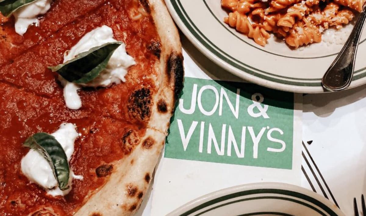 A close-up of a margherita pizza with basil and cheese sits next to creamy pasta on a table with a green "Jon & Vinny's" sign—sometimes listed among famous pizzerias not worth visiting.