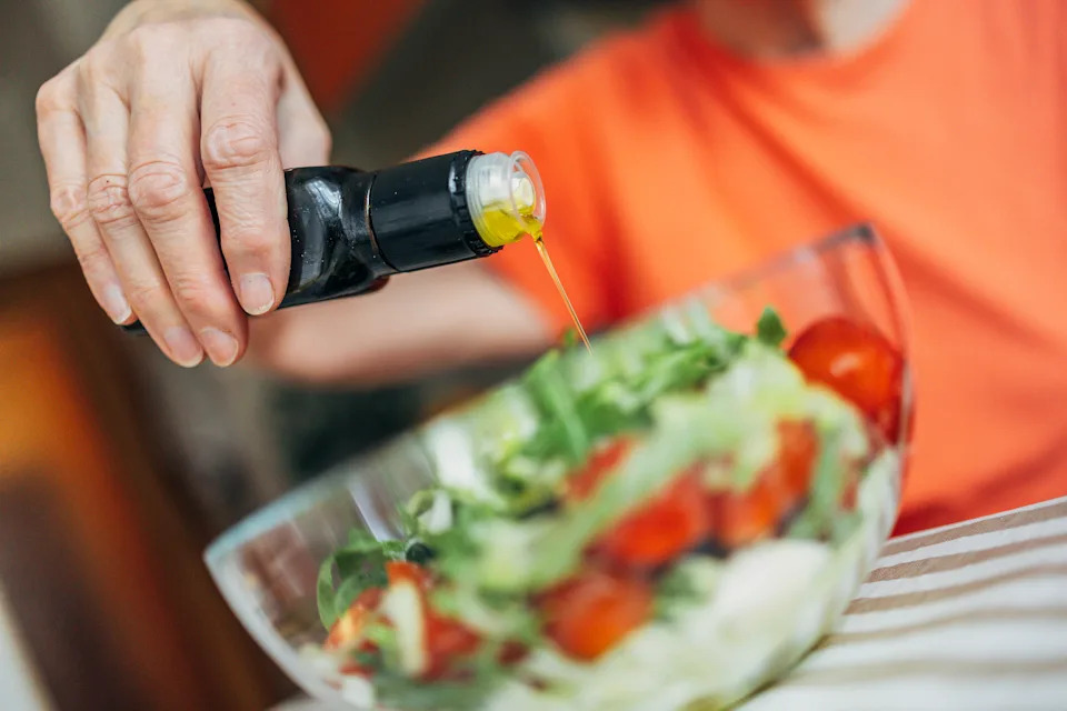 Older adult hand pouring olive oil on fresh garden salad.