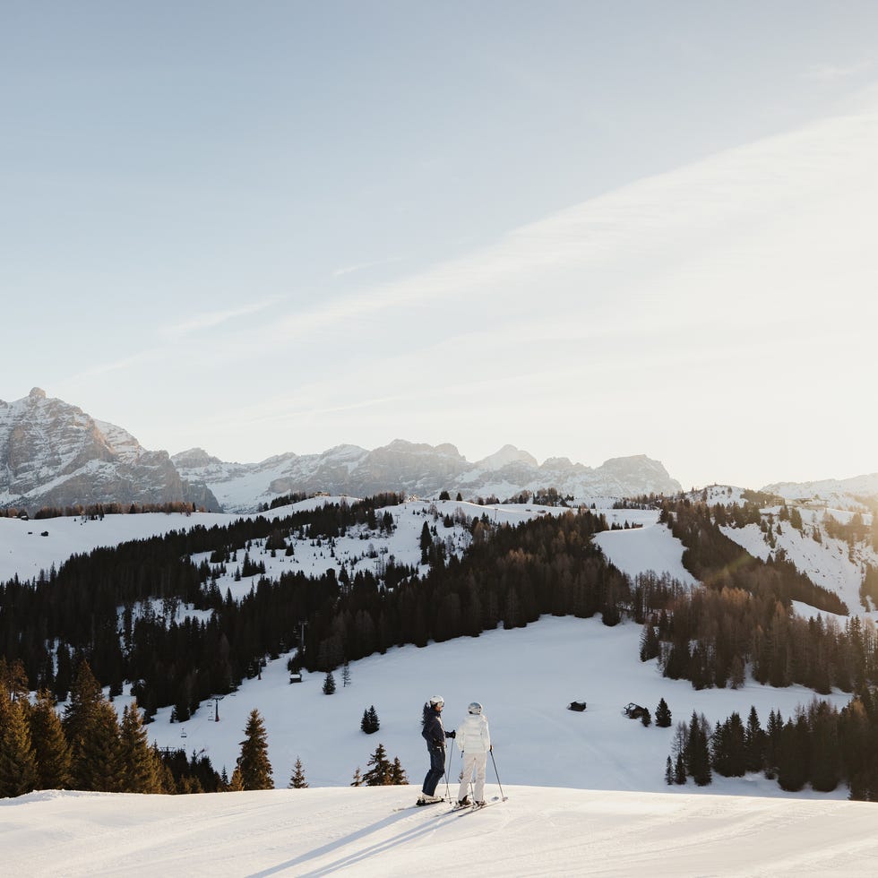 two skiers overlooking a snowy landscape with mountains in the background during sunset two skiers overlooking a snowy landscape with mountains in the background during sunset