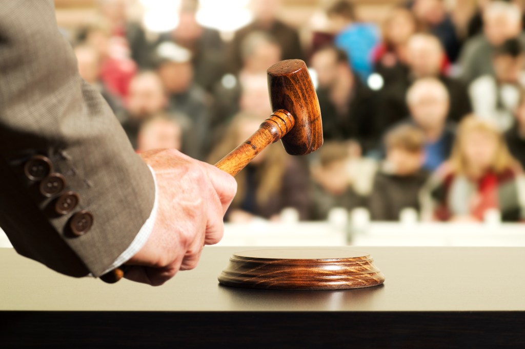 A hand holding a judge's gavel over a base with a blurred crowd in the background.