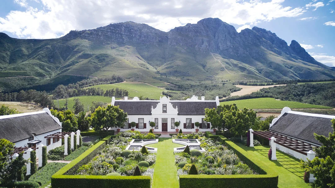 A large, white Cape Dutch-style manor with thatched roofs sits in a manicured garden, surrounded by green lawns and hedges, with dramatic mountains rising in the background under a partly cloudy sky.