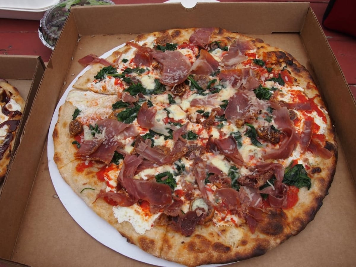 A freshly baked pizza in an open cardboard box, topped with prosciutto, spinach, melted cheese, tomato sauce, and a crispy, slightly charred crust.