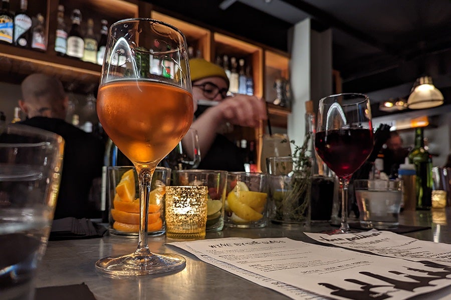 Wine glasses sit on a silver bar in a dimly lit space, with bottles lining illuminated wooden shelves on the backbar.