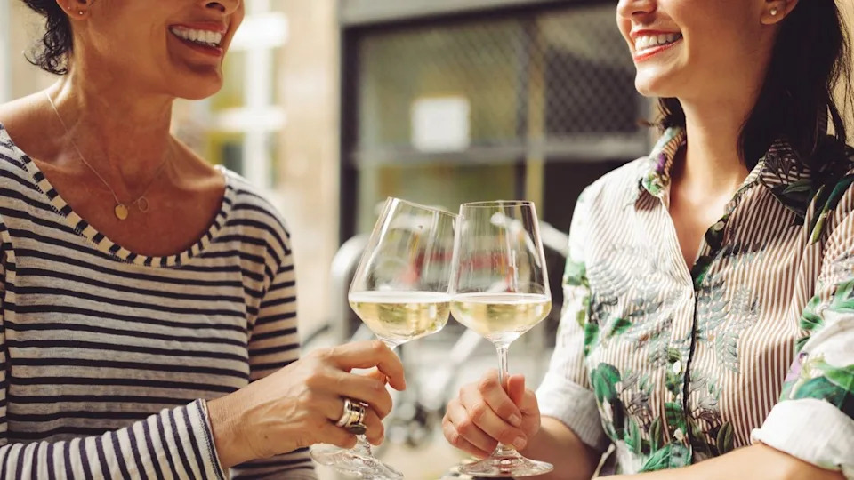  Two women smiling as they clink their respective glasses of white wine together . 