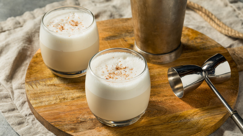 Two cream colored milk punch cocktails with a dusting of nutmeg on a round wooden serving board.