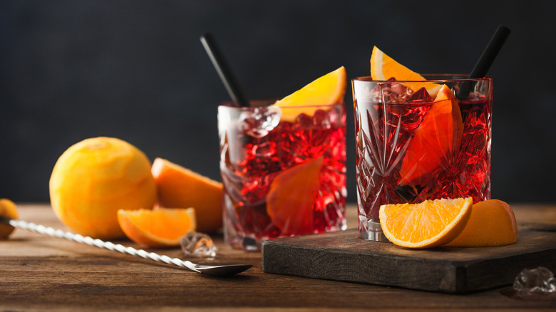Two Negronis on a wooden board surrounded by fresh orange wedges.