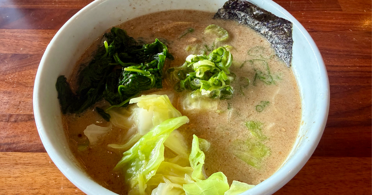 Bowl of creamy broth with green vegetables, including spinach, leafy greens, and sliced cabbage, garnished with green onions and a piece of seaweed, placed on a wooden table.