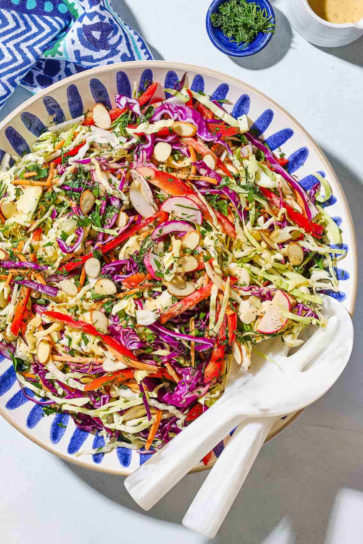 An overhead photo of cabbage salad in a serving bowl with serving utensils. Next to this is a kitchen towel, a bowl of dill, and a small container of the dressing.