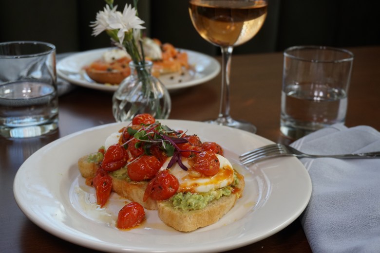 Avocado toast with poached egg, roasted cherry tomatoes, and microgreens served at a restaurant table with rosé wine.