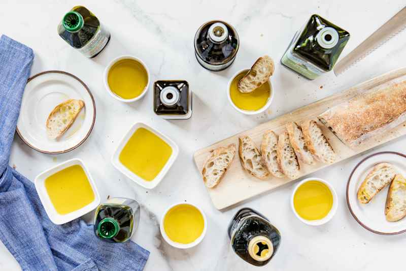 Various olive oil bottles and ramekins filled with olive oil on table with sliced bread on cutting board.