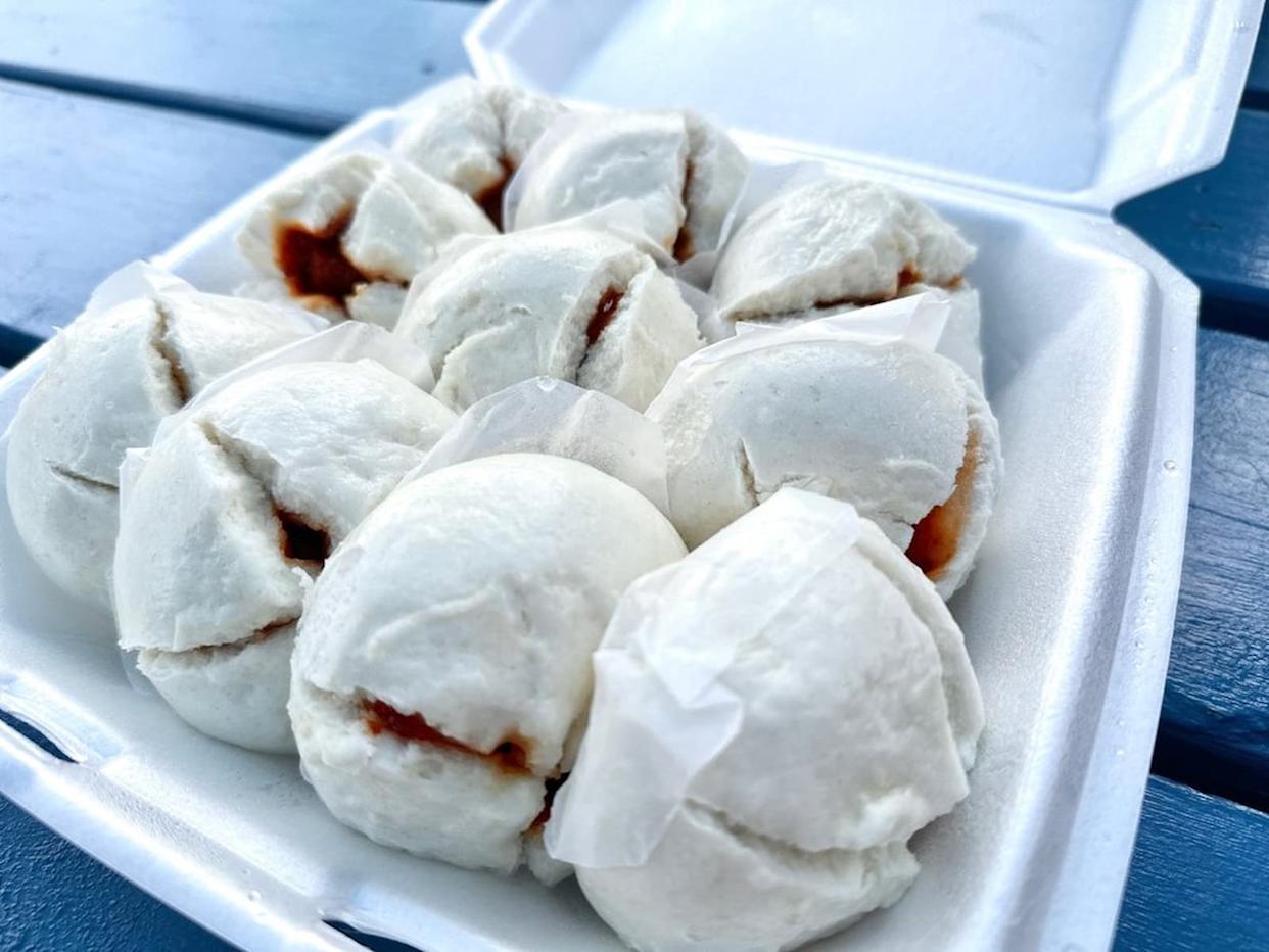 A white takeout container holds nine steamed pork buns with cracked tops, revealing a hint of brown filling inside. The buns are placed on a blue table, and some have small pieces of parchment attached.