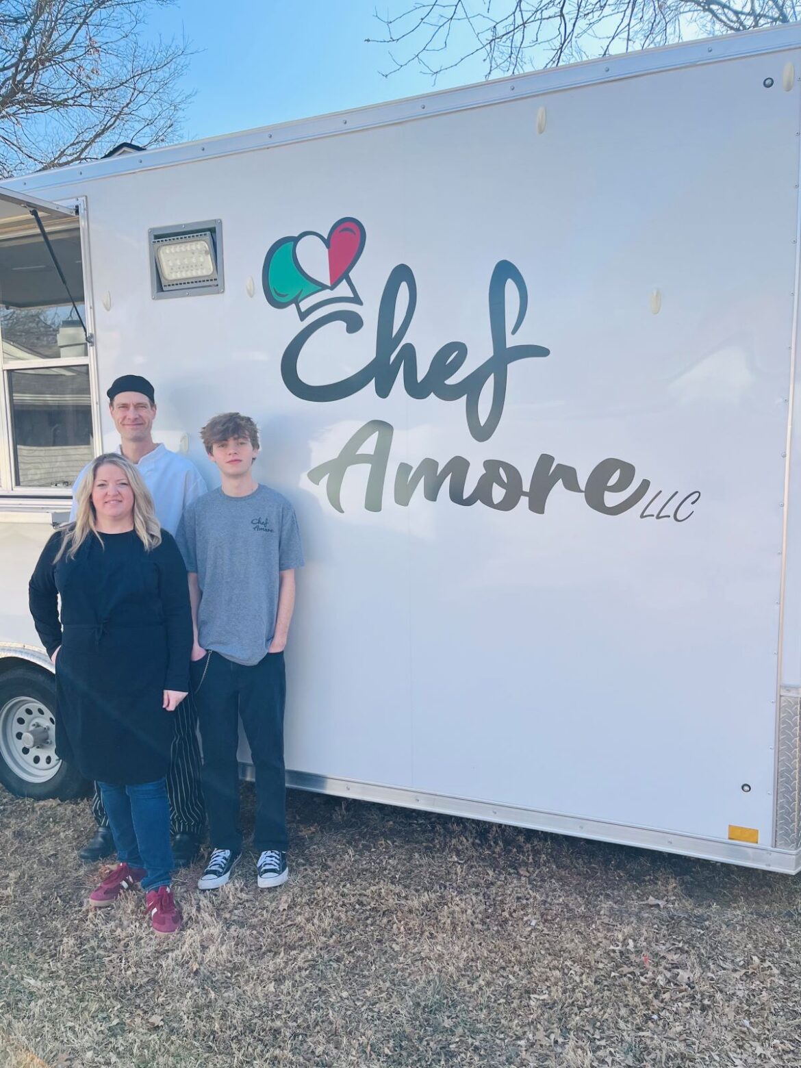 Three people stand outdoors beside a white food truck trailer with the logo “Chef Amore LLC” displayed in large lettering. A woman wearing a black apron stands in front, with a man in a chef’s jacket and hat and a teenage boy in a gray T-shirt standing behind her. The photo appears to be taken on a sunny day in a grassy yard with trees nearby.