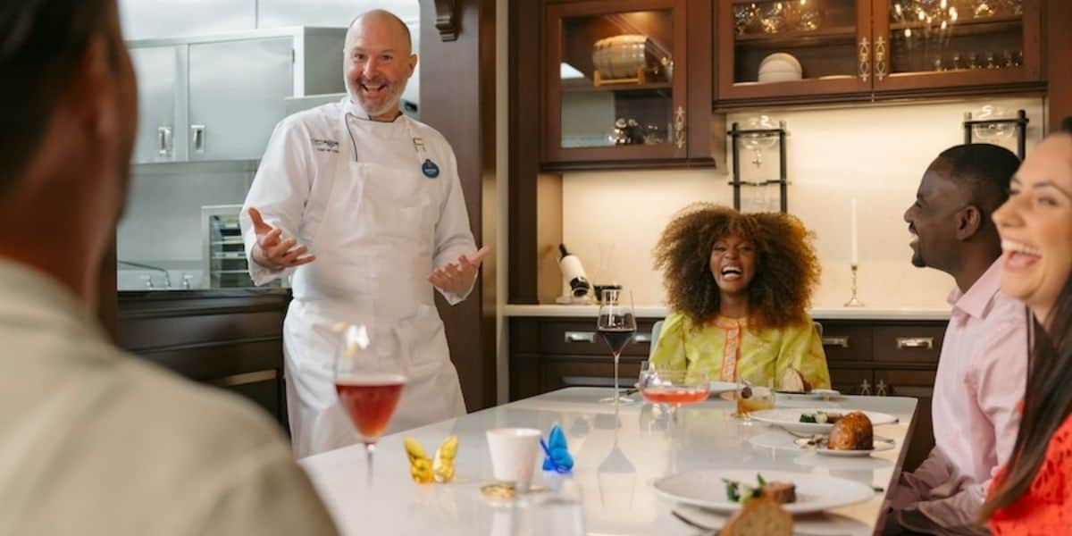 A chef in a white uniform is laughing and gesturing while speaking to four people seated around a table at the Chef's Table at Victoria & Albert's.