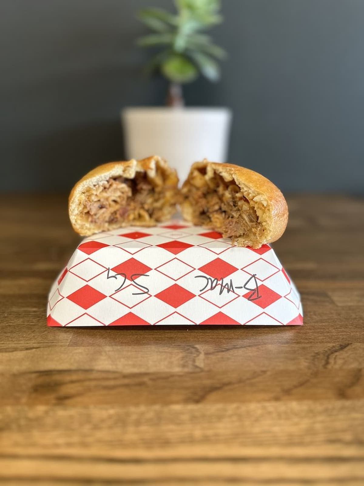 A golden-brown stuffed bun, cut in half to reveal shredded meat filling, sits on a red-and-white checkered paper atop a wooden table; a small potted plant is blurred in the background.