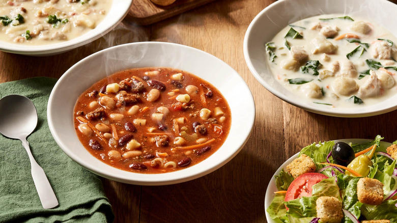 White bowls of various soups from Olive Garden next to bowl of salad on wooden table