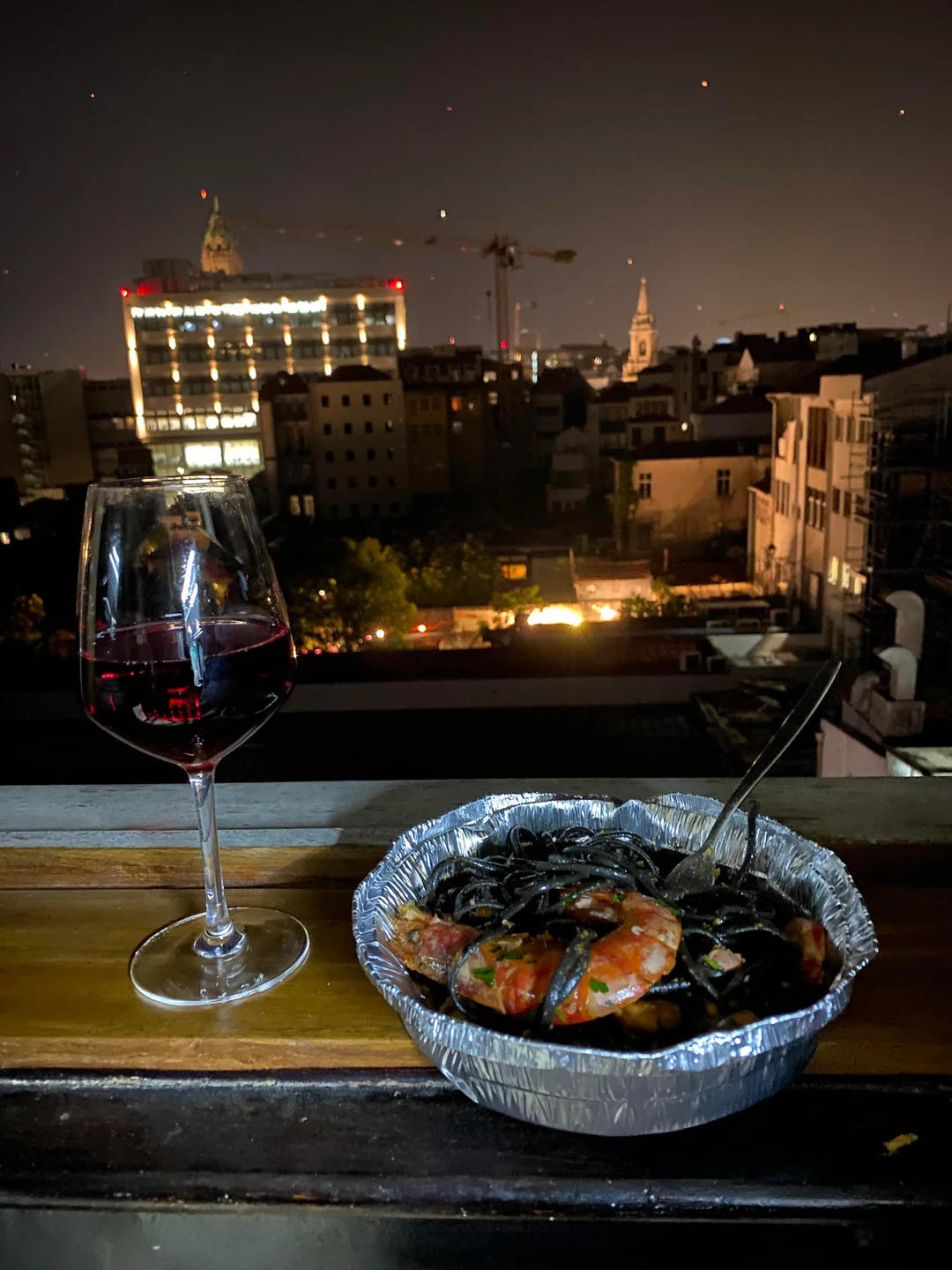 A glass of red wine and a foil container of seafood pasta are set on a ledge, with a nighttime city skyline featuring lit buildings and cranes in the background.