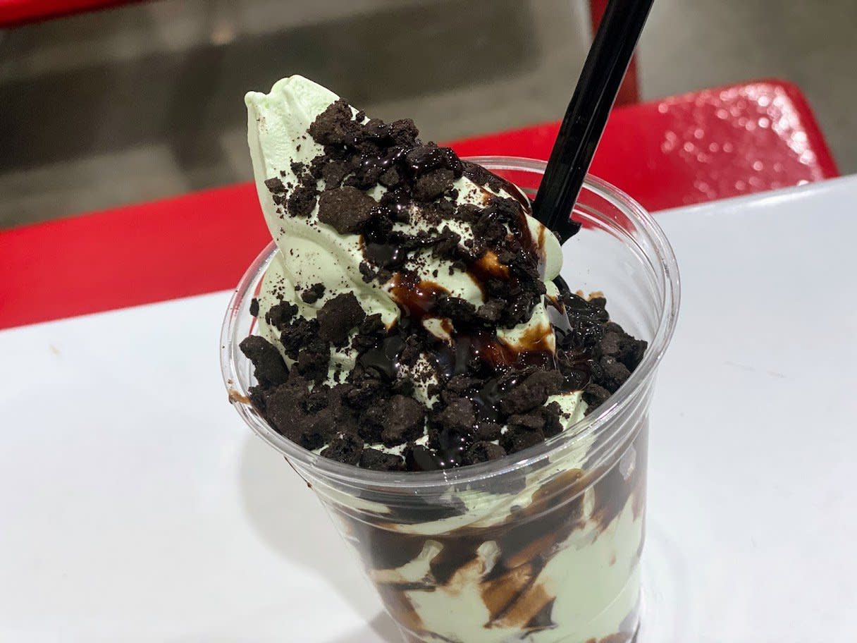 A cup of mint green soft-serve ice cream topped with chocolate sauce and crushed cookies, with a black spoon inserted, sits on a white table with a red bench in the background.