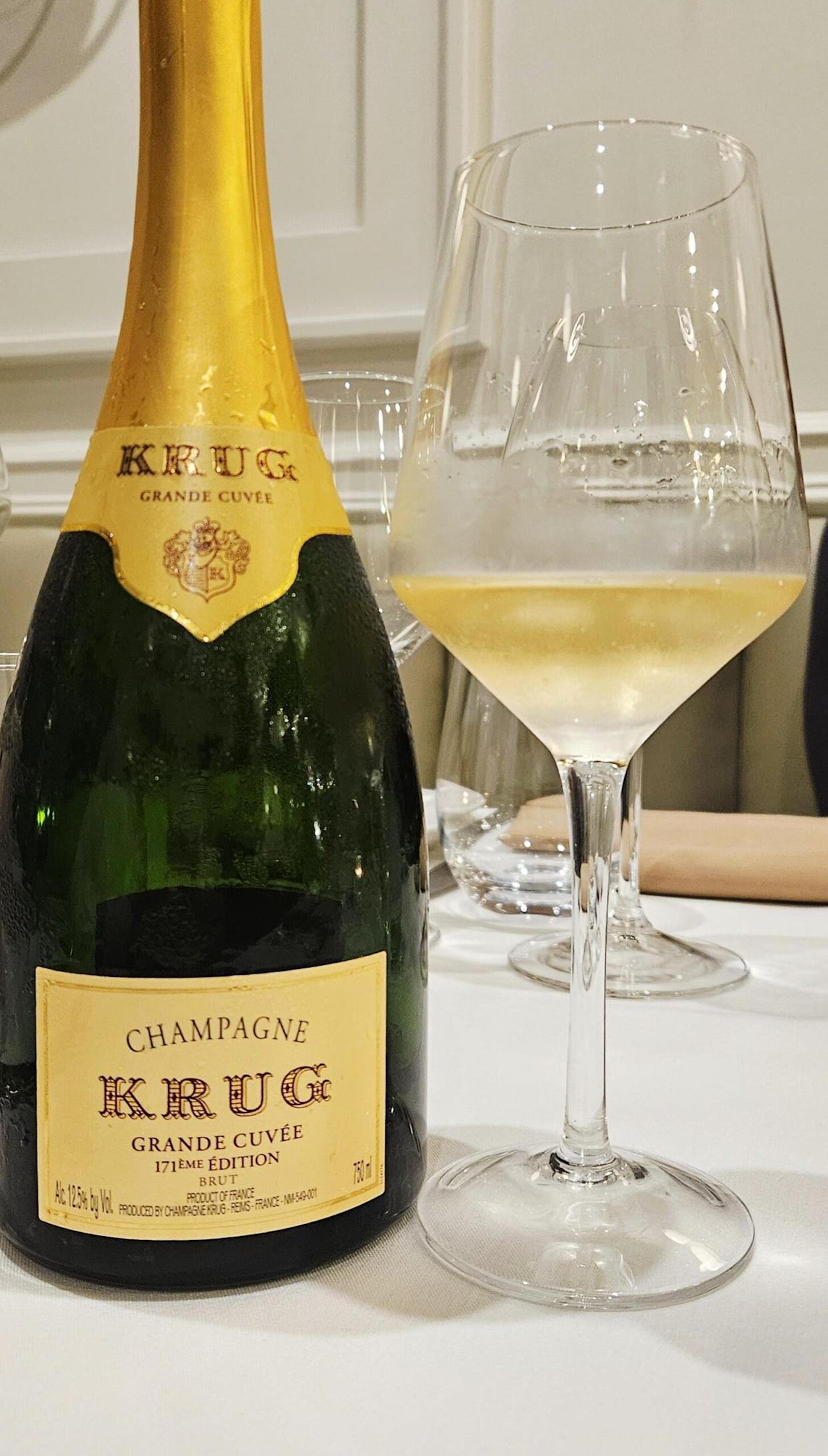 A bottle of Krug Grande Cuvée Champagne is placed on a white tablecloth next to a filled wine glass. The background shows more glassware and a neutral, elegant setting.