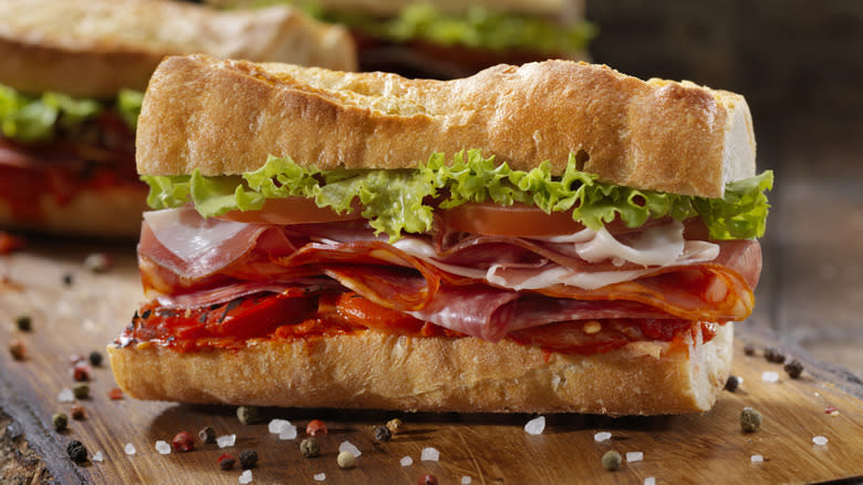 An Italian sub sits on a wooden board surrounded by salt and pepper