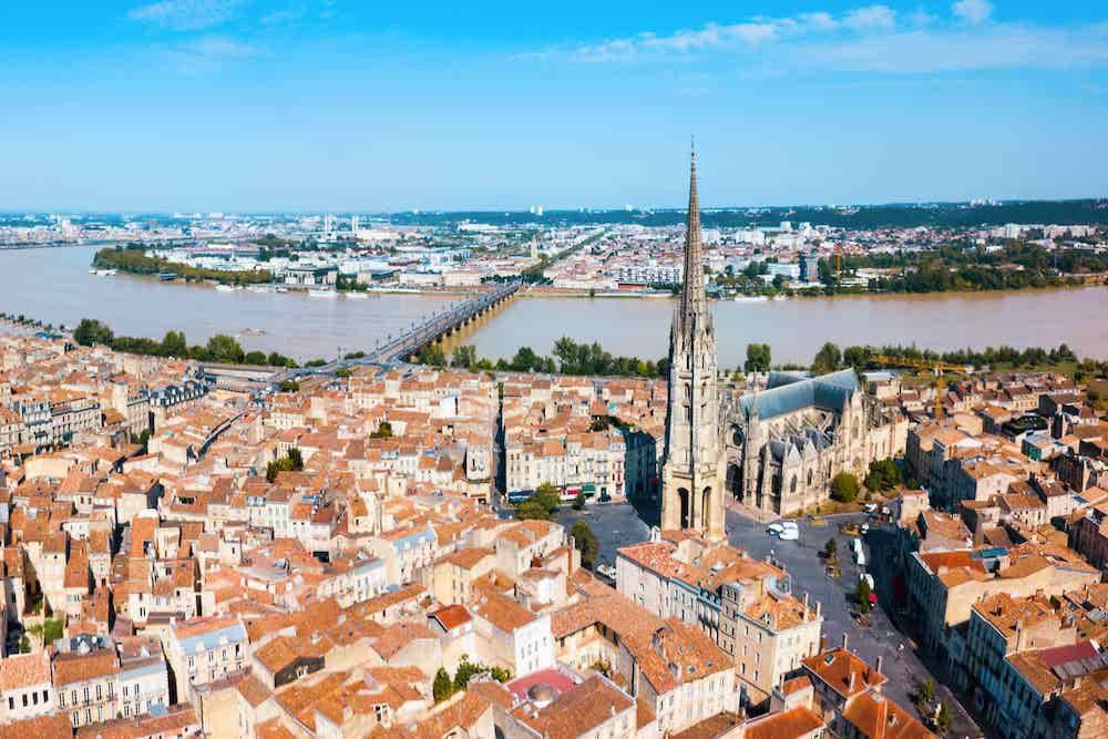 Aerial view of a historic European city with red-tiled roofs, a large Gothic church with a tall spire in the center, and a wide river with bridges in the background under a clear blue sky.