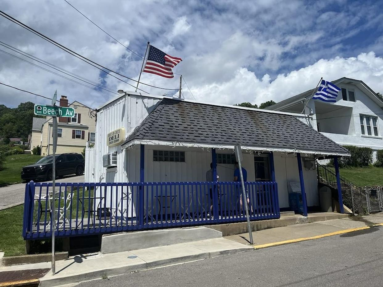 A small white building with blue trim and a ramp, flying American and Greek flags. A street sign says "Beech AV." Two people stand near the entrance under a partly cloudy sky.