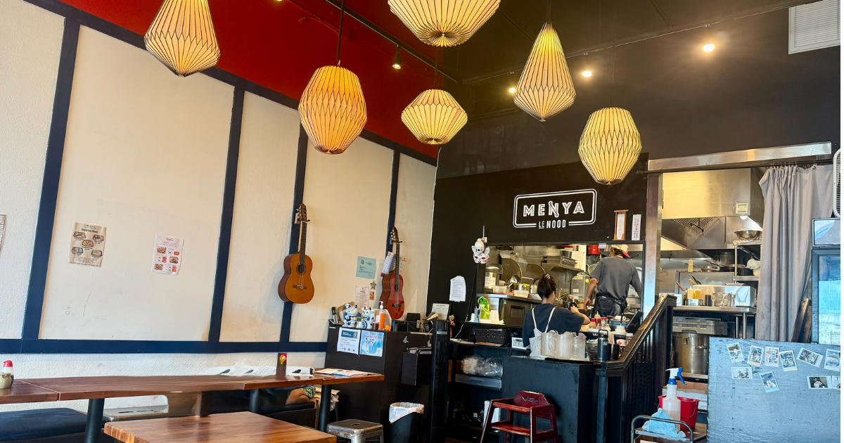 Interior of a cozy restaurant featuring decorative hanging lanterns, wooden tables, and a visible kitchen area with staff at work.