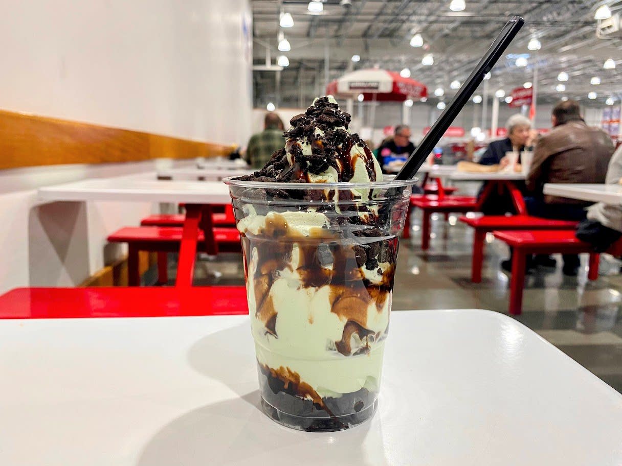 A clear plastic cup filled with vanilla ice cream, chocolate sauce, and crumbled cookies sits on a white table in a casual dining area with red benches and people in the background. A black spoon is in the cup.