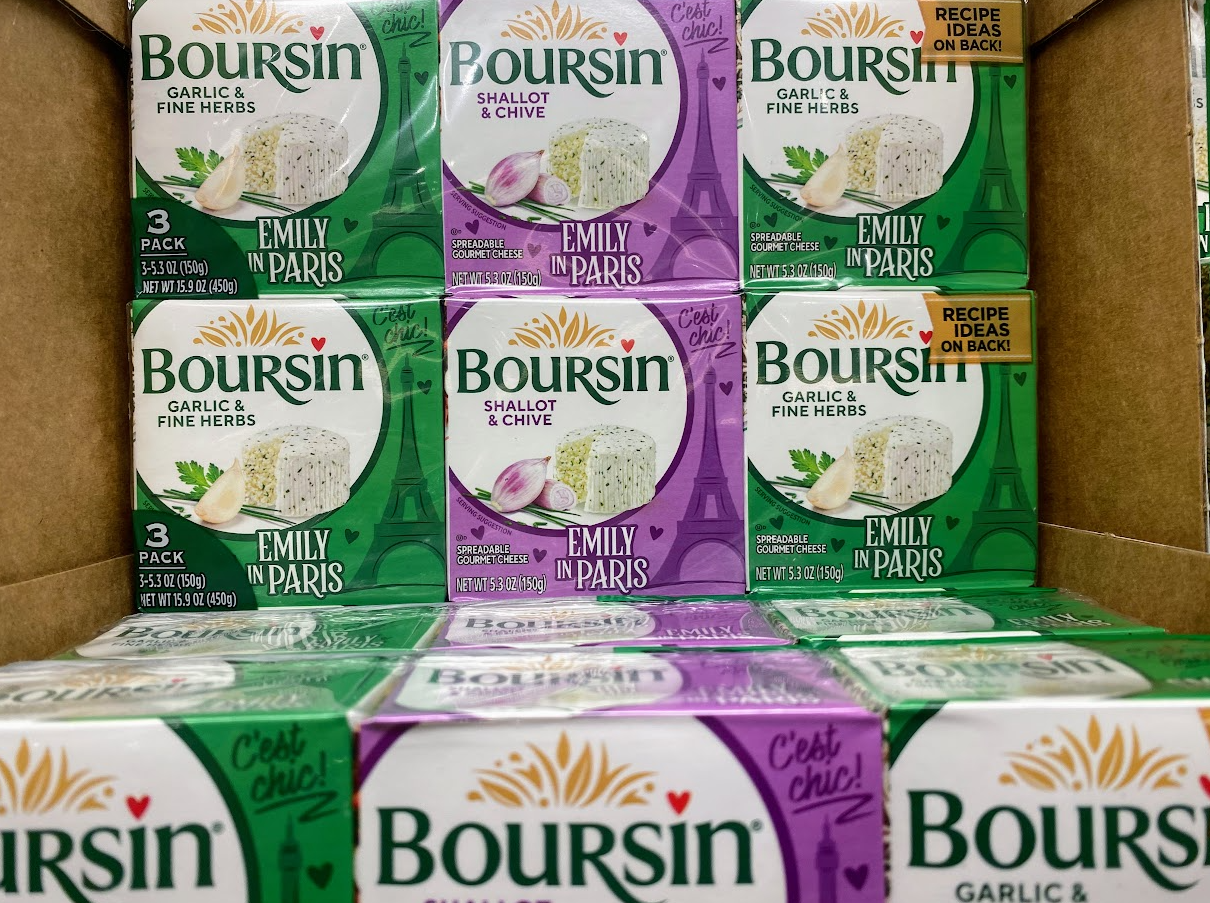 Boxes of Boursin cheese, including Garlic & Fine Herbs and Shallot & Chive flavors, are stacked in a display box. Packaging features "Emily in Paris" branding and a note about recipe ideas on the back.