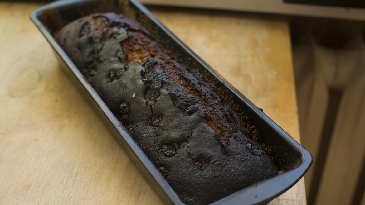 A burnt loaf of bread sits in a black baking pan on a wooden surface.