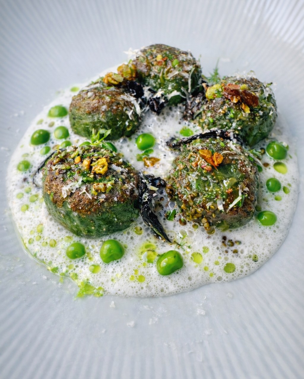 five balls of green gnudi from the los angeles restaurant orsa & winston sit in a foamy white sauce made with dashi and celery root cream. the dish is topped with cheese and green peas can be seen in the sauce.