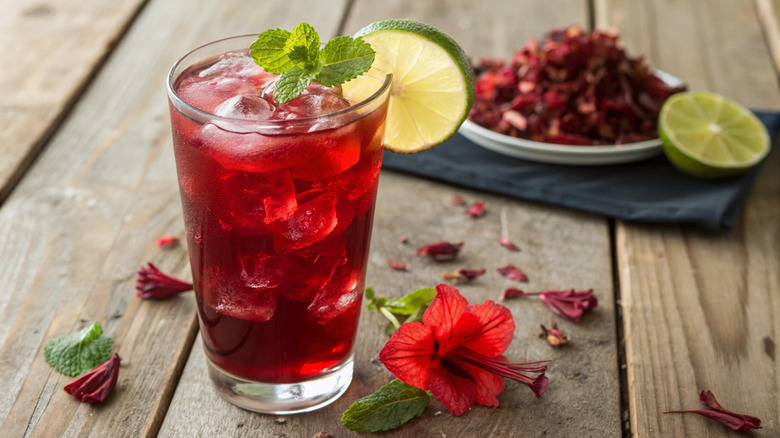 A tall glass of vibrant red iced tea with ice, lime, and mint sits on a rustic wooden surface with scattered hibiscus petals.
