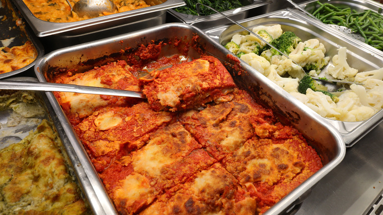 A buffet tray of lasagna is seen next to trays of steamed vegetables