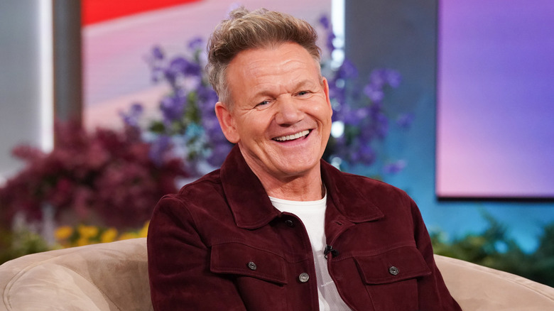 Celebrity chef Gordon Ramsay sits laughing in a dark red shirt in a beige armchair.