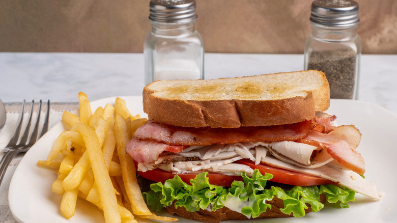 A BLT sandwich on toasted white bread with sliced turkey on plate with side of french fries