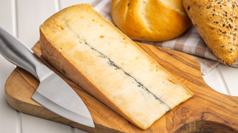 A large wedge of Morbier on a cutting board next to a cheese knife and loaves of bread