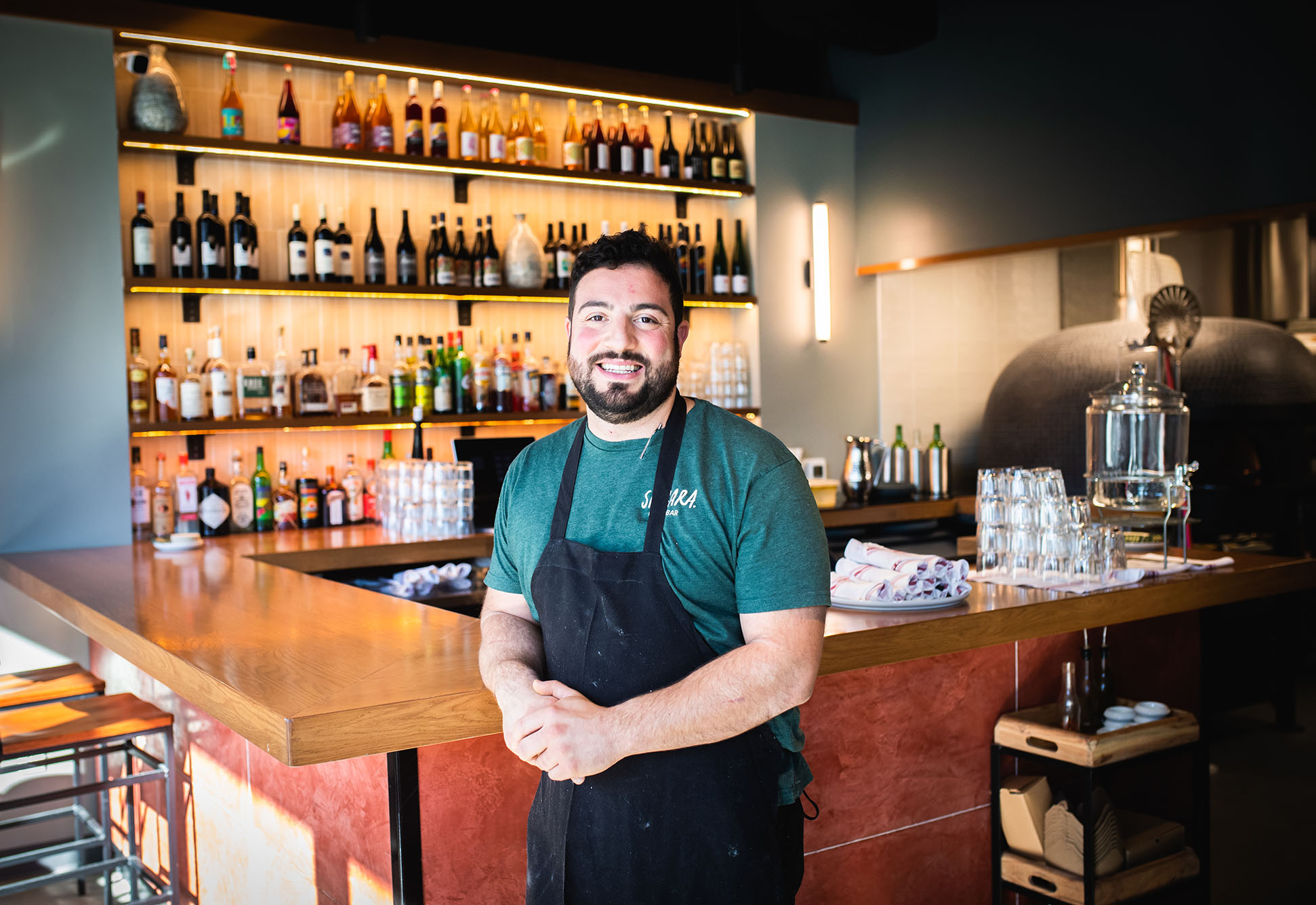 Photo: Michael Lombardi, Jr. (Questrom'08) poses at Si Cara, a neighborhood pizzabar in Cambridge on November 9, 2022. A white man with dark brown beard and haircut smiles and poses in front of a wooden bar. He wears a green shirt and black apron and leans back against wooden counter. Wooden shelves in back feature a variety of alcoholic drinks.